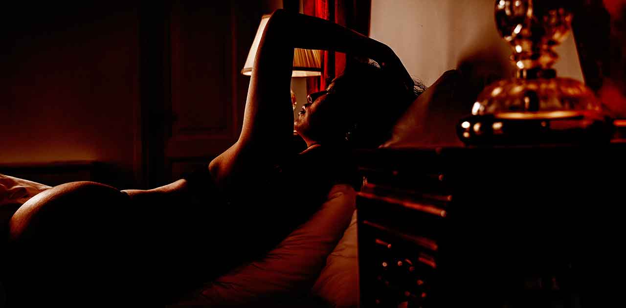 Boudoir foto van een vrouw in zwarte lingerie. Ze licht op bed in een sierlijke pose. Foto heeft veel warme kleuren.