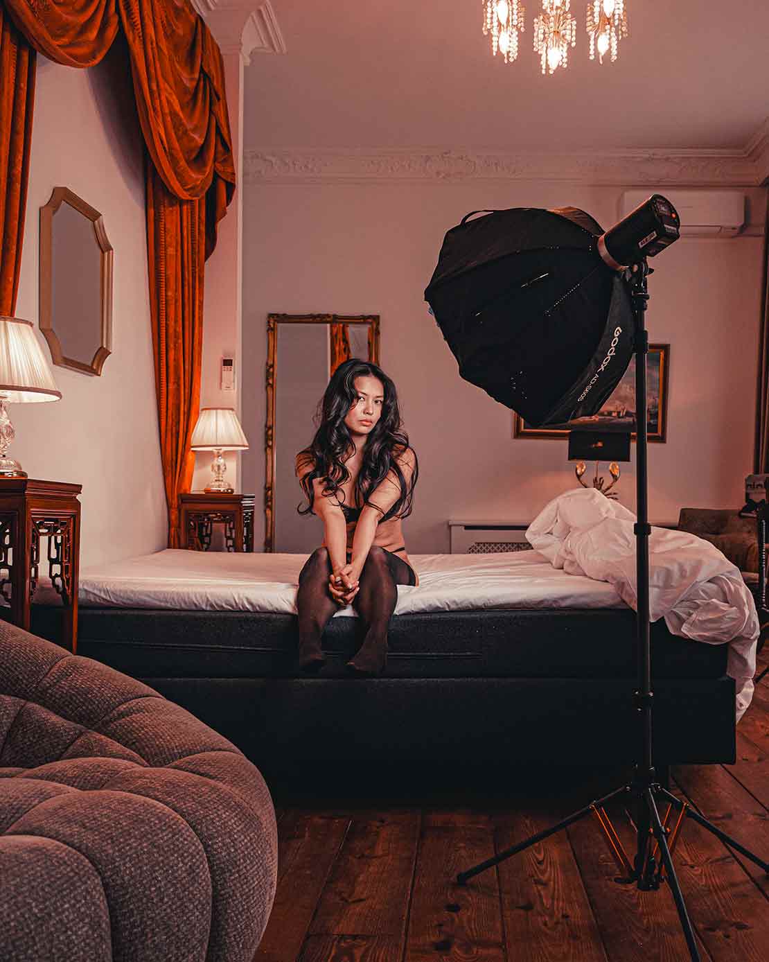 Boudoirfoto van een vrouw in lingerie, zittend bij het raam in een lichte slaapkamer.