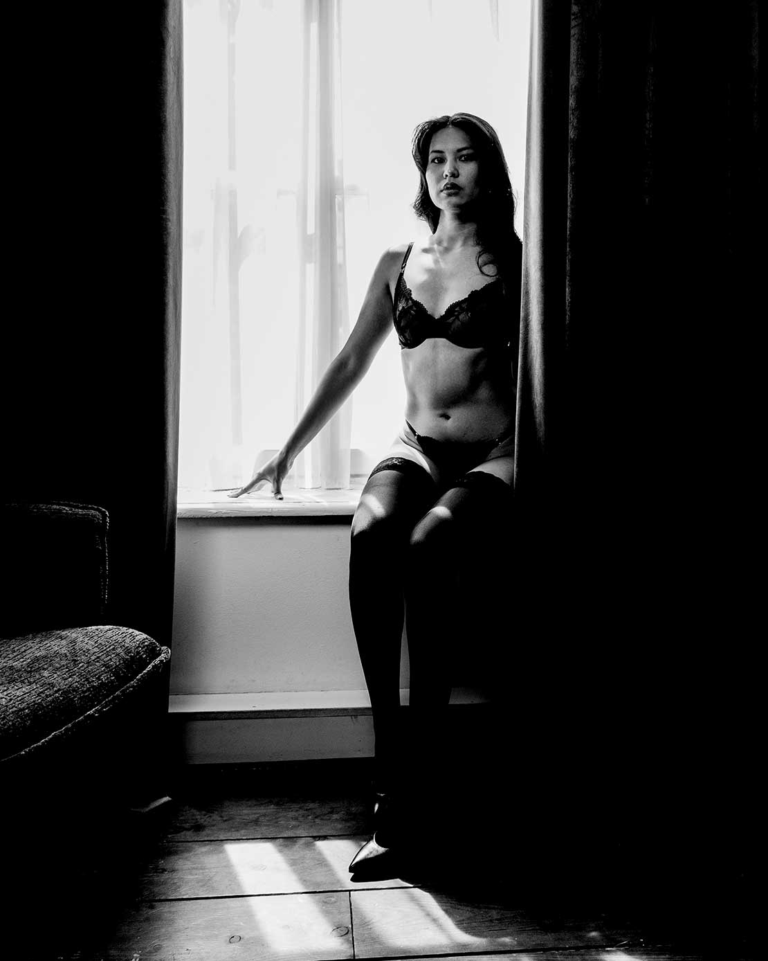 Boudoirfoto van een vrouw zittend bij een groot raam in een sfeervolle slaapkamer.