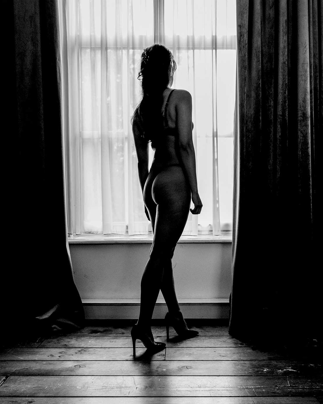 Boudoirfoto van een vrouwelijk silhouet voor een groot raam in een sfeervolle slaapkamer.