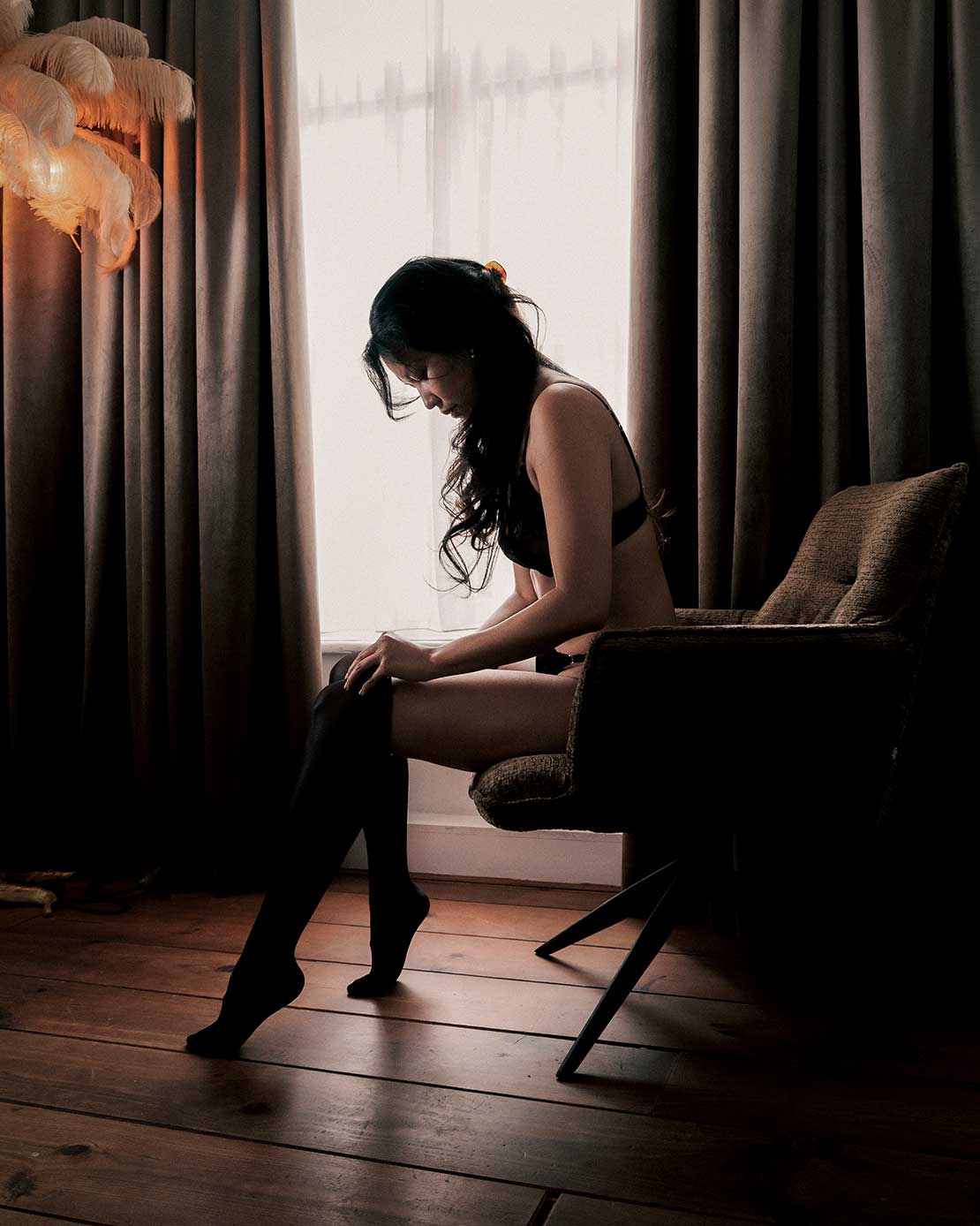Boudoirfoto van een vrouw zittend op een stoel bij het raam in een sfeervolle kamer.