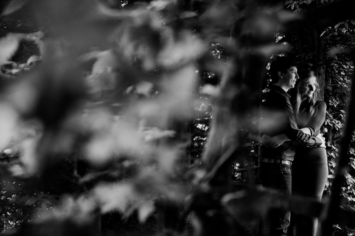 Foto van Jeroen Koeten Fotografie. Loveshoot gemaakt in Drenthe. Foto is in zwart-wit. In de foto kijk je door de bosjes naar ze.